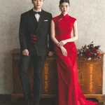 Full body couple wedding portrait, bride in red long train wedding cheongsam (qipao) leaning on vintage wooden cabinet, classic chinese bridal dress for wedding ceremony