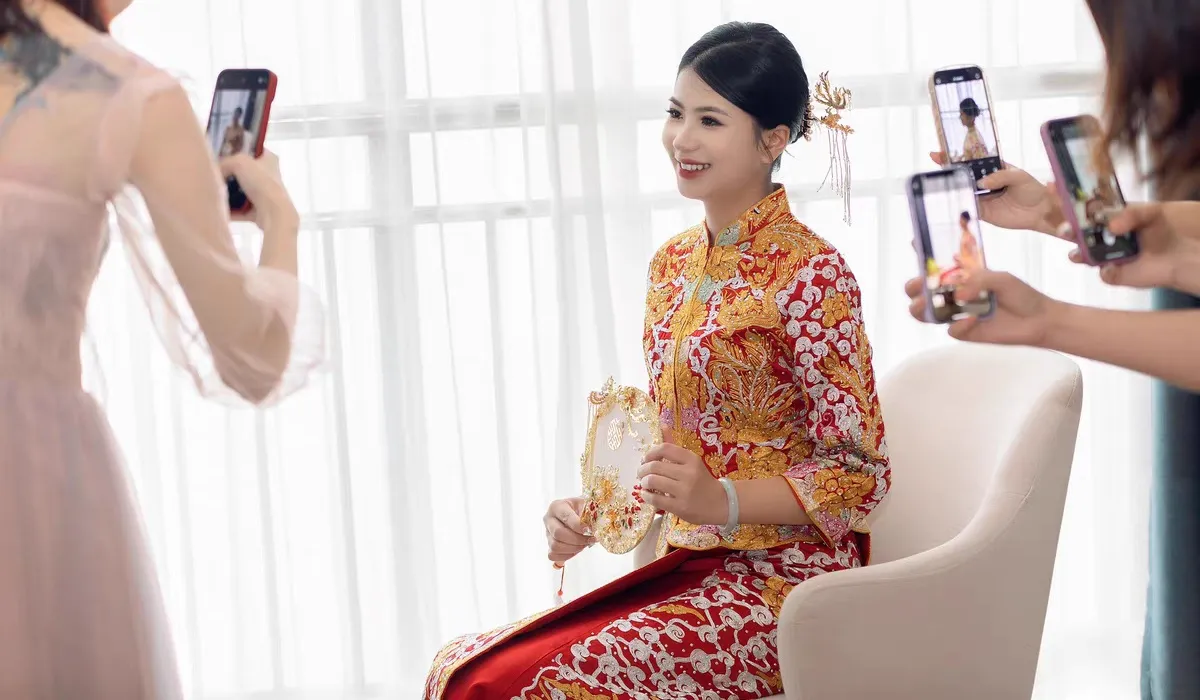  Beautiful Chinese bride wearing a traditional Qun Kwa bridal dress, holding a decorative fan while friends capture her wedding look with smartphones.