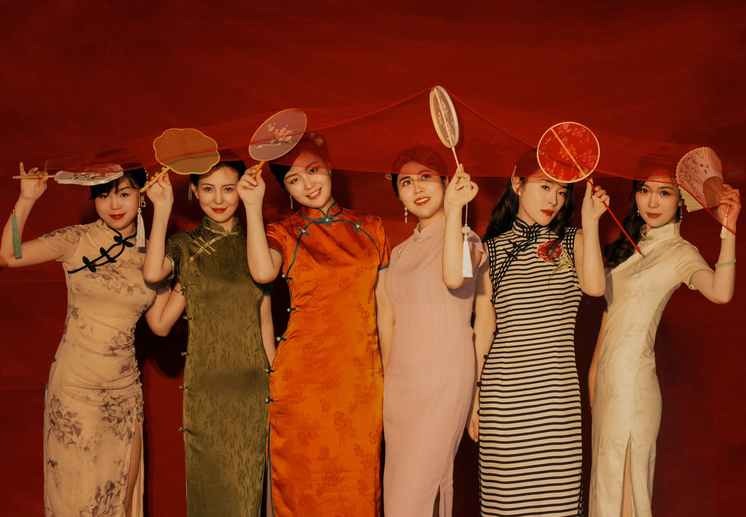 Vintage-style group portrait of women in classic traditional cheongsam qipao dresses against a red background, featuring various designs like floral and solid color qipao, ideal for learning what a cheongsam is and its timeless aesthetic.