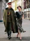 Woman in a 1930s long qipao of dark green brown mulberry silk walking side by side with a man in a military cape, highlighting the dress's fluid abstract print and high slit in a vintage city street backdrop.