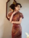 Woman standing in front of a wooden-framed mirror, wearing a 1930s reddish-brown floral silk qipao, with her reflection visible to highlight the dress's smooth silk texture and intricate floral pattern from a side angle.