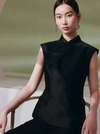Model wearing a black cheongsam top, women's sleeveless qipao style mandarin collar top, styled with black trousers while sitting in a soft-lit minimalist studio.