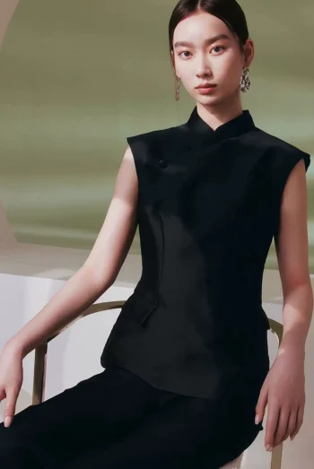 Model wearing a black cheongsam top, women's sleeveless qipao style mandarin collar top, styled with black trousers while sitting in a soft-lit minimalist studio.