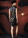 Back view of model in black silk mini cheongsam dress, showcasing the tailored back silhouette, all-over koi fish jacquard pattern, and smooth premium silk fabric in a modern studio setting.
