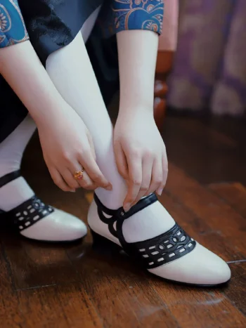 Black and white 1920s Qipao dress goat leather shoes with classic hollow out details, worn with white socks, a model adjusts the shoes on wooden floor, ideal for vintage cheongsam styling.