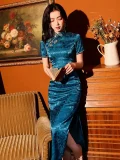 Woman holding a vintage book while wearing a luxurious blue silk long qipao dress with high slit, showcasing the soft drape of the floral jacquard fabric under warm studio lighting.