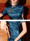 Close-up detail shot of a blue silk qipao dress, focusing on the mandarin collar, delicate frog buttons, and intricate floral jacquard texture on the short sleeves.