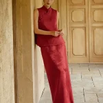 Two-piece Burgundy Modified Qipao With Pants (or Matching Skirt) - Woman in elegant burgundy qipao set with sleeveless mandarin collar brocade top and coordinating maxi skirt, leaning against a stone wall in a sunlit classic corridor, modern chinese dress qipao for formal events and weddings