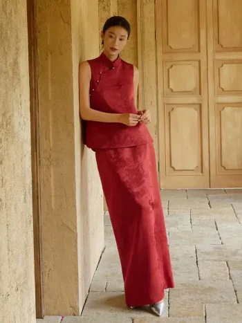Two-piece Burgundy Modified Qipao With Pants (or Matching Skirt) - Woman in elegant burgundy qipao set with sleeveless mandarin collar brocade top and coordinating maxi skirt, leaning against a stone wall in a sunlit classic corridor, modern chinese dress qipao for formal events and weddings