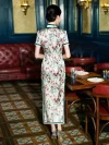 Back view of a model in a Chinese silk qipao dress with white floral print, standing in a vintage restaurant to show the tailored back design and full-length silhouette.
