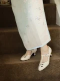 Close-up of cream womens vintage high heel qipao sandals with genuine leather T strap design, worn with a light blue floral cheongsam on carpeted stairs, showcasing intricate cutout details and retro charm