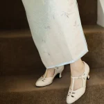 Close-up of cream womens vintage high heel qipao sandals with genuine leather T strap design, worn with a light blue floral cheongsam on carpeted stairs, showcasing intricate cutout details and retro charm