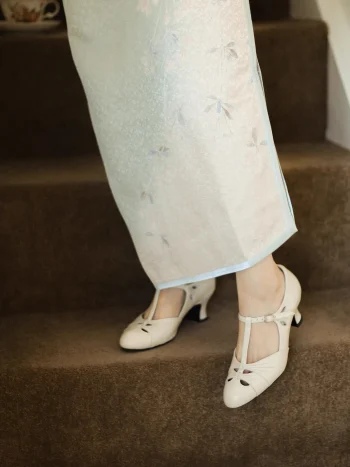 Close-up of cream womens vintage high heel qipao sandals with genuine leather T strap design, worn with a light blue floral cheongsam on carpeted stairs, showcasing intricate cutout details and retro charm