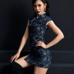 Woman model seated on dark steps, wearing a dark blue peacock print short cheongsam dress with silver trim and high side slit, highlighting the tailored fit and modern style of this fashion cheongsam.