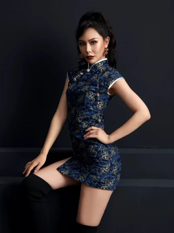 Woman model seated on dark steps, wearing a dark blue peacock print short cheongsam dress with silver trim and high side slit, highlighting the tailored fit and modern style of this fashion cheongsam.