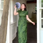 Plus size model wearing dark green floral print chinese qipao dress, elegant form-fitting cheongsam with traditional mandarin collar, posing with a folding fan in a bright doorway setting.