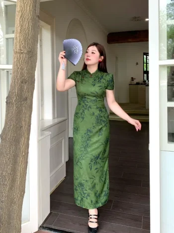 Plus size model wearing dark green floral print chinese qipao dress, elegant form-fitting cheongsam with traditional mandarin collar, posing with a folding fan in a bright doorway setting.