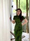 Plus size woman in dark green floral print chinese qipao dress, elegant traditional cheongsam styled with a folding fan, posing on a wooden patio with greenery in the background.