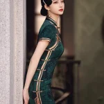Elegant woman in dark green vintage floral qipao with traditional frog buttons, leaning against a stone pillar, showcasing the side slit and retro silhouette.