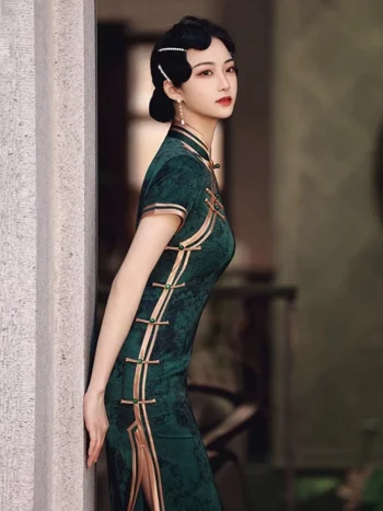 Elegant woman in dark green vintage floral qipao with traditional frog buttons, leaning against a stone pillar, showcasing the side slit and retro silhouette.