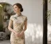 Elegant non-Chinese woman respectfully wearing a cream floral qipao with green trim, posing in a classical Chinese garden with bamboo and moon gate, showcasing cultural appreciation of traditional Chinese fashion.