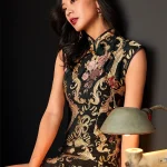 Elegant woman seated in a studio, wearing a fashion black and gold embroidered short qipao dress with intricate floral and baroque-style embroidery, showcasing the mandarin collar, tassel details, and luxurious texture of this black embroidered qipao.