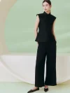 Full-body view of a model wearing a women's black cheongsam top, a sleeveless qipao style mandarin collar top, styled with black wide-leg trousers in a minimalist studio with soft green background.