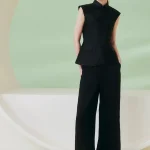 Full-body view of a model wearing a women's black cheongsam top, a sleeveless qipao style mandarin collar top, styled with black wide-leg trousers in a minimalist studio with soft green background.