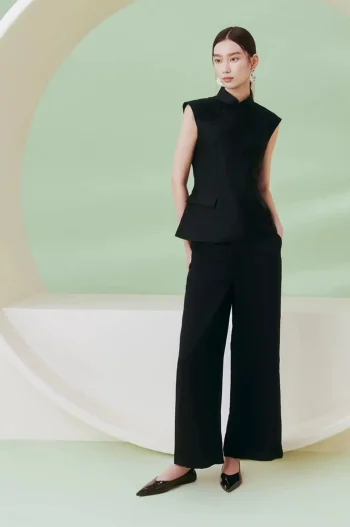 Full-body view of a model wearing a women's black cheongsam top, a sleeveless qipao style mandarin collar top, styled with black wide-leg trousers in a minimalist studio with soft green background.