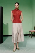 Full-body view of a model wearing a women's red chinese slim fit jacquard cheongsam top, a red modern mandarin collar cheongsam top, styled with a light jacquard midi skirt and gold shoes.