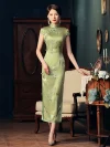 Full-body shot of a woman wearing a green slim fit long cheongsam dress, standing in front of a vintage cabinet, showing the traditional frog buttons and floral print details.