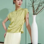Model wearing a green modern chinese mulberry silk cheongsam shirt, women's short sleeve qipao top with water drop collar, paired with a light skirt in a minimalist interior setting.