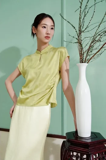 Model wearing a green modern chinese mulberry silk cheongsam shirt, women's short sleeve qipao top with water drop collar, paired with a light skirt in a minimalist interior setting.
