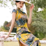 Model seated on a white birch fence, wearing a women high end yellow mulberry silk cheongsam dress with traditional Chinese landscape print, showcasing the elegant fit and satin sheen.