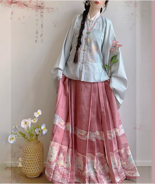Woman wearing light blue square-collar Ming Dynasty aoqun with pink horse-face skirt, showcasing delicate embroidery and intricate landscape patterns, styled in a soft traditional Chinese studio with floral decor.