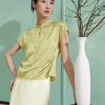 Side view of a woman wearing a light green mulberry silk modern cheongsam shirt, featuring a flattering ruched waist detail that enhances the silhouette of this elegant short sleeve qipao top.