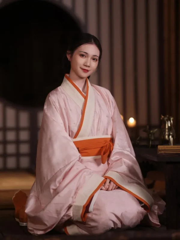 Woman wearing soft light pink quju hanfu, an elegant ancient Chinese robe with contrasting orange trim, sitting gracefully in a dimly lit traditional wooden interior with warm ambient lighting.