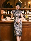 Stylish woman in a misty blue bamboo print brocade cheongsam dress paired with a vintage birdcage veil hat, leaning against a wooden bar to highlight the dress's elegant lines and high slit.