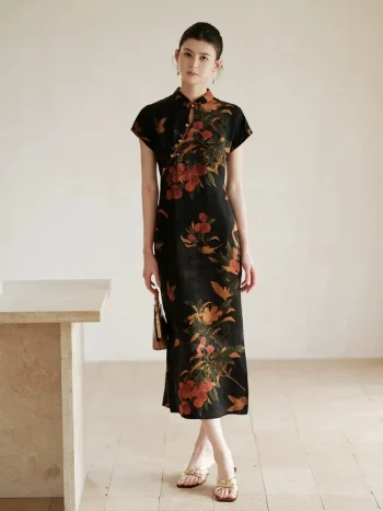 Full length view of a modern Chinese qipao dress, a slim fit black mulberry silk cheongsam with vibrant lychee and floral print. The elegant woman stands in a bright, minimalist room, highlighting the dress's tailored silhouette.