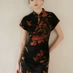Model leaning against a soft beige wall, wearing a modern high end lightweight short sleeve cheongsam dress. The black mulberry silk qipao features a rich nature inspired print, perfect for elegant everyday wear.
