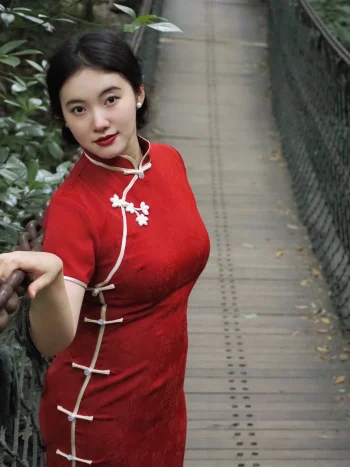 Plus size model posing on a forest suspension bridge in a modern red midi qipao, featuring delicate white floral frog buttons and a tailored silhouette. This contemporary red qipao blends classic Chinese details with everyday wearability.