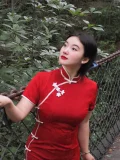 Half-body portrait of a woman in a red modern midi qipao, leaning on a bridge chain and looking upward in a lush forest. The qipao's subtle embossed fabric and elegant fit shine in natural light.