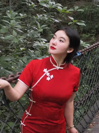 Half-body portrait of a woman in a red modern midi qipao, leaning on a bridge chain and looking upward in a lush forest. The qipao's subtle embossed fabric and elegant fit shine in natural light.