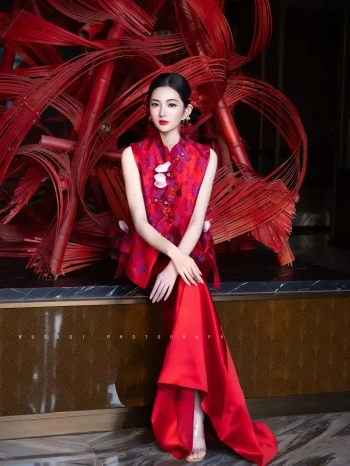Modern Red Silk Qipao Set - Full-body shot of woman in two-piece red qipao set, featuring a sleeveless brocade top and flowing satin skirt, sitting in front of a red bamboo art installation, ideal modern red qipao for formal occasions