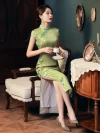 Woman sitting on a vintage chair in a mulberry silk green cheongsam dress, holding a teacup and saucer, showcasing the dress's high slit and tailored fit in a classic interior setting.