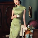 Elegant woman holding a white teacup, wearing a mulberry silk green slim fit long cheongsam dress with magnolia print, posing in a vintage-style room to show the smooth fabric and traditional mandarin collar.