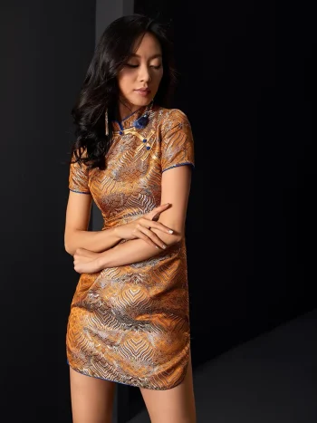 Woman wearing orange gold brocade jacquard short qipao dress with blue trim, form-fitting modern chinese qipao for cocktail parties, crossed arms pose in dark studio.