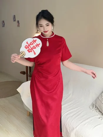 Plus size model wearing a vibrant red satin qipao dress, elegant chinese red qipao with pearl-trimmed mandarin collar and keyhole neckline, posing with a wedding fan in a warm modern home setting.