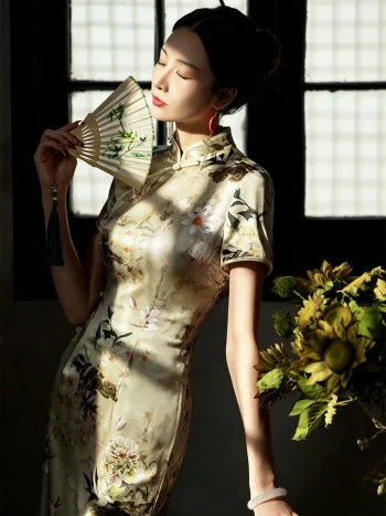 Model wearing a premium 19 momme mulberry silk printed midi qipao, holding a bamboo folding fan by a sunlit window, showcasing the elegant floral print and lustrous satin texture.