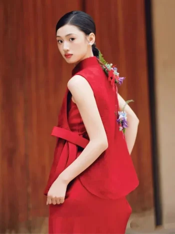 Red Chinese Cheongsam Dress - Back view of bride in 2 piece cheongsam, showcasing the sleeveless mandarin collar vest's back design with a ribbon tie, standing in front of a wooden door with floral hair accessories, modern red cheongsam for weddings.